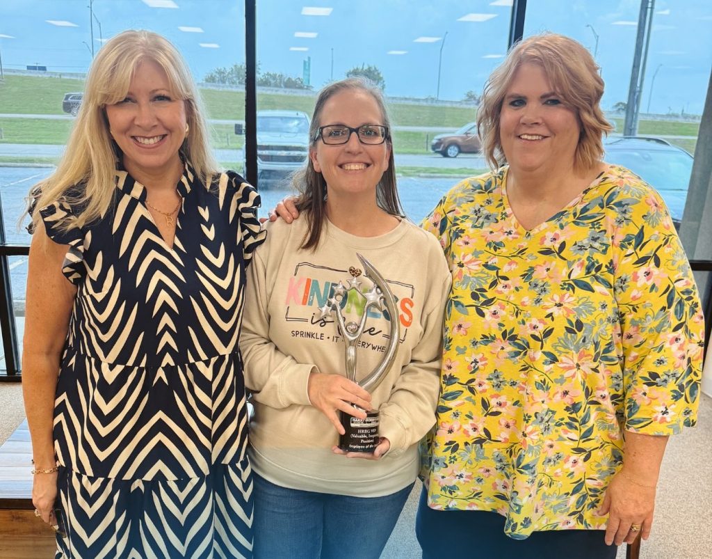 Three women standing together, smiling and posing for a photo. The woman in the middle holds an award trophy. They are in an indoor setting with windows showing a green landscape outside.